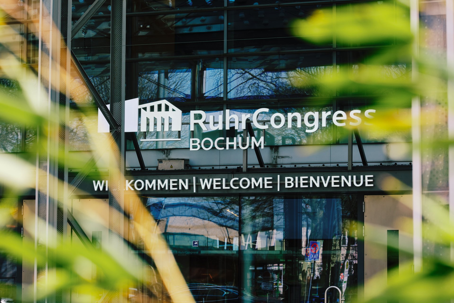 Close-up of the RuhrCongress Bochum logo at the main entrance with green bamboo leaves in the foreground.
