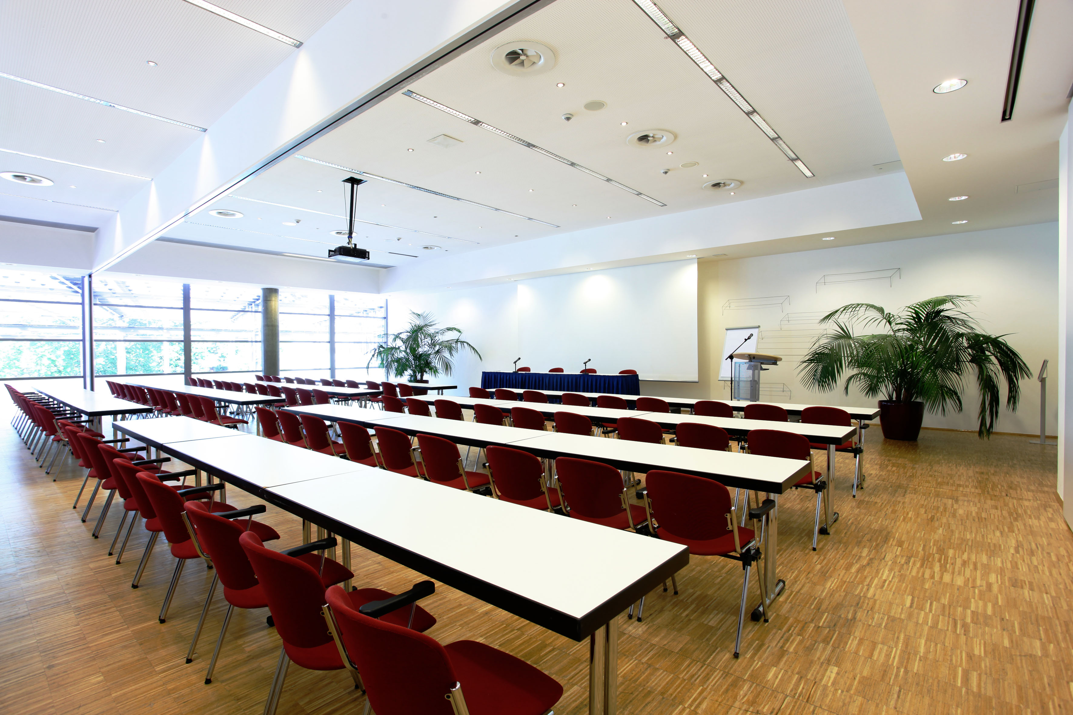 Parlamentarische Bestuhlung im Tagungsraum im RuhrCongress Bochum. Im Hintergrund hängt eine große Leinwand, davor steht ein Plenumstisch, sowie ein Rednerpult. Links und rechts steht jeweils eine große grüne Pflanze.